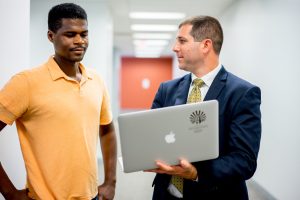 Two men in hallway conversing, one with a computer