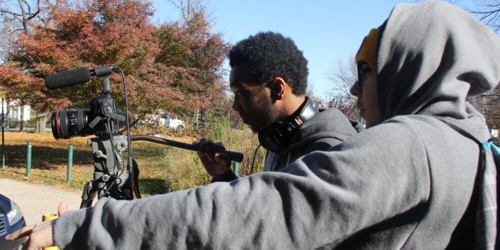 Rox 3 one student is looking through a video camera while another student is providing film direction beside him