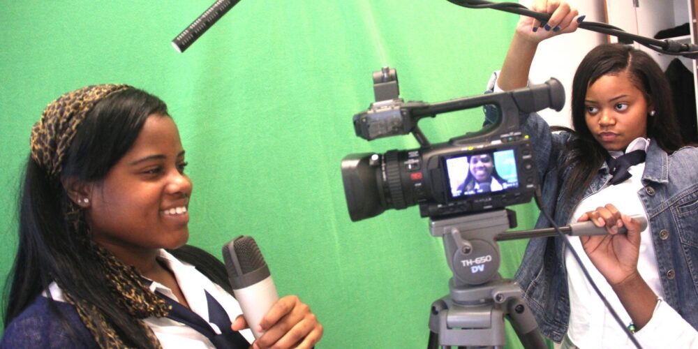 a behind the scenes preview of video production. A student holding a microphone stands in front of a green screen while two other students are handling a video camera and an overhead microphone