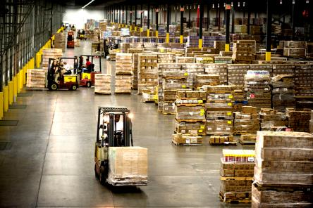 the inside of a warehouse filled with crates and boxes. Forklifts carry large boxes along the main hallway
