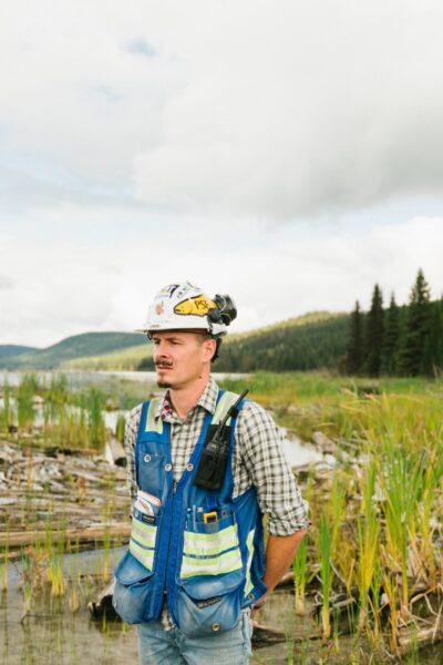pexels-mount-polley-3580281 a man wearing a safety vest and hard hat stands in the middle of a marsh, inspecting the land before him