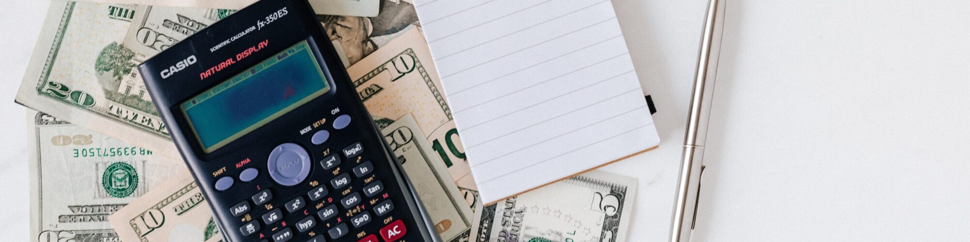 a calculator and a small notebook lays on top of a pile of cash on a table