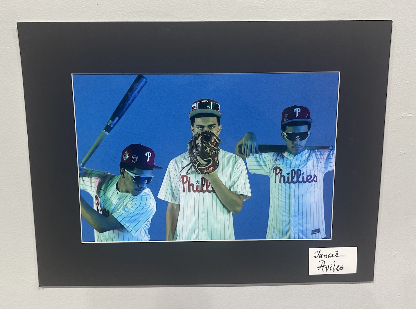 Artwork by student Janiah Aviles of a baseball player depicted three times over a blue background. On left, posed to hit a ball. Middle, holding glove in front of face to only show eyes. On right, holding baseball bat over shoulders with both hands.