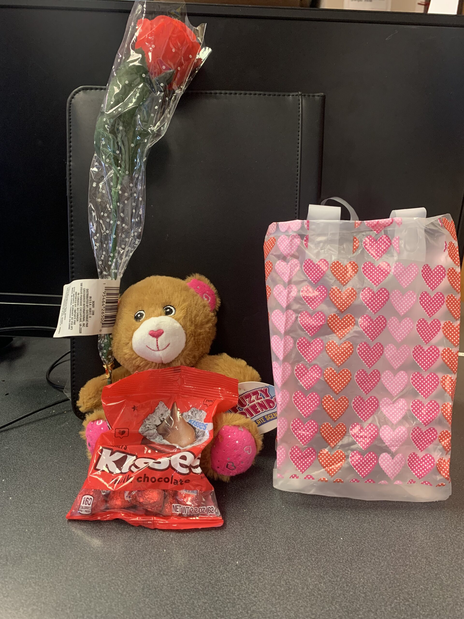 a brown teddy bear, a small bag of Hershey Valentine's Kisses, and a faux Rose, next to matching Valentine's Day gift bag.