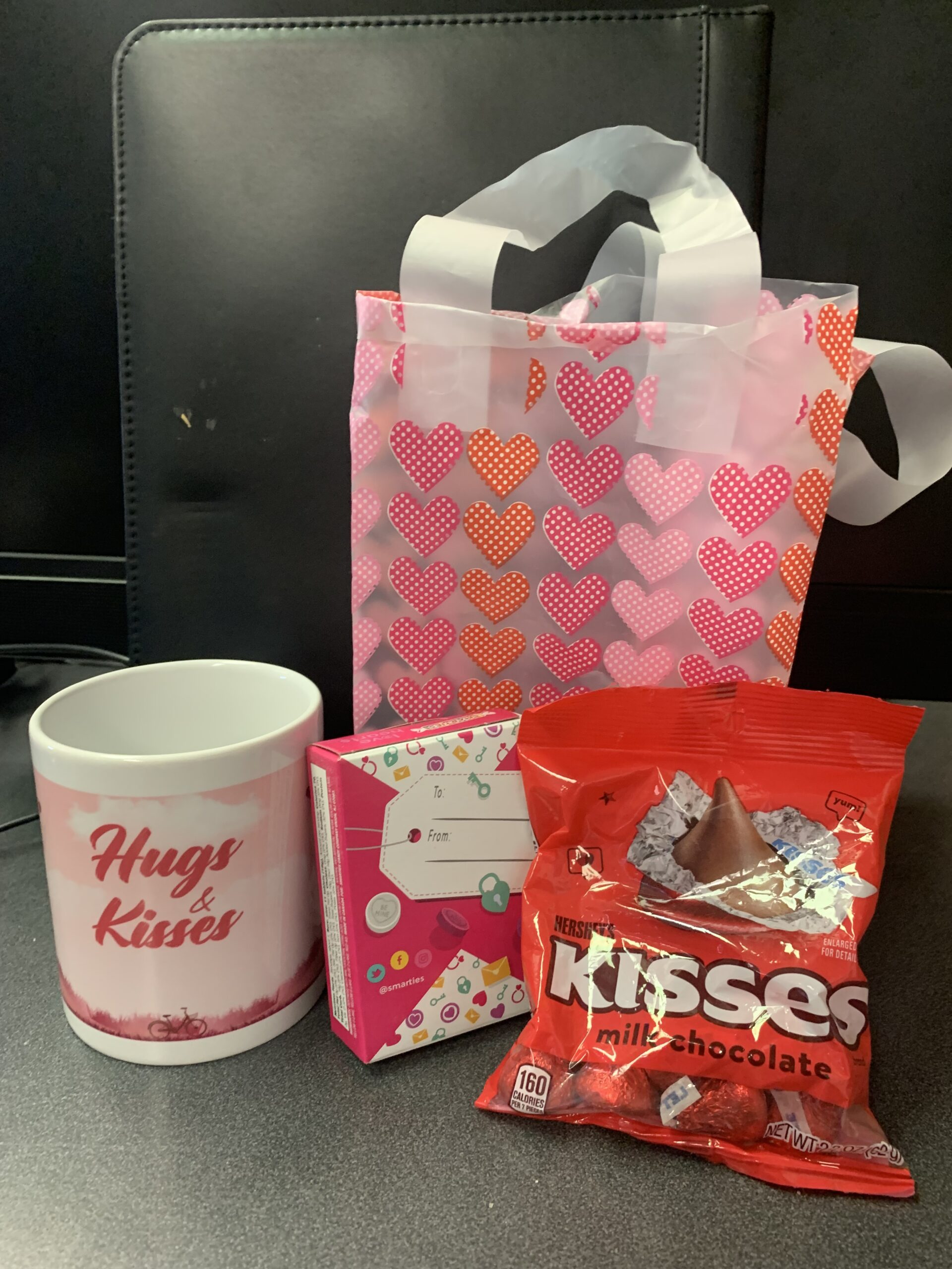 A small ceramic mug with print saying "Hugs & Kisses", a small box of heart candies, and a small bag of Hershey's Valentine's Kisses, in front of a matching Valentine's Day gift bag.