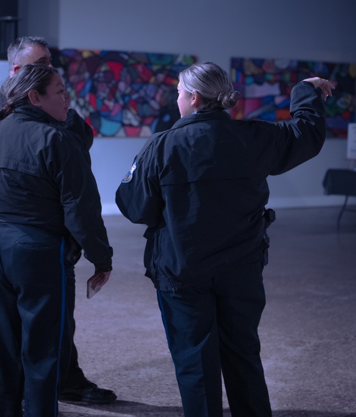 A candid photo taken during KCAPA's "Story Time" art exhibit. A group of three police officers are conversing with their backs turned to the camera. The background shows two artworks that look like mosaics.