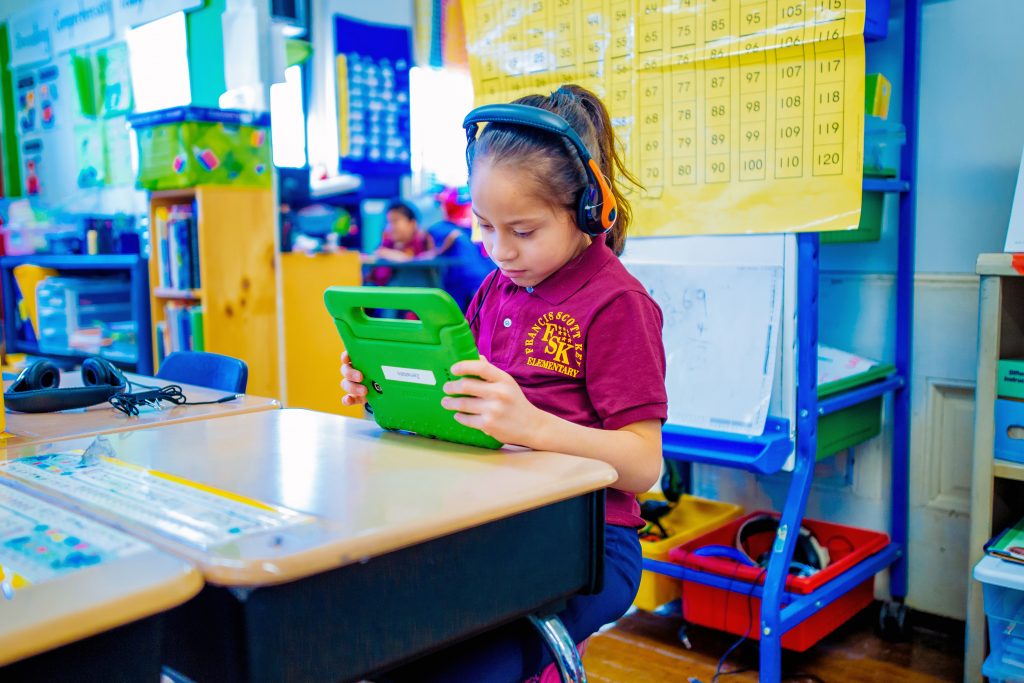 Image of an elementary school girl learning on an iPad with headphones on at her desk.