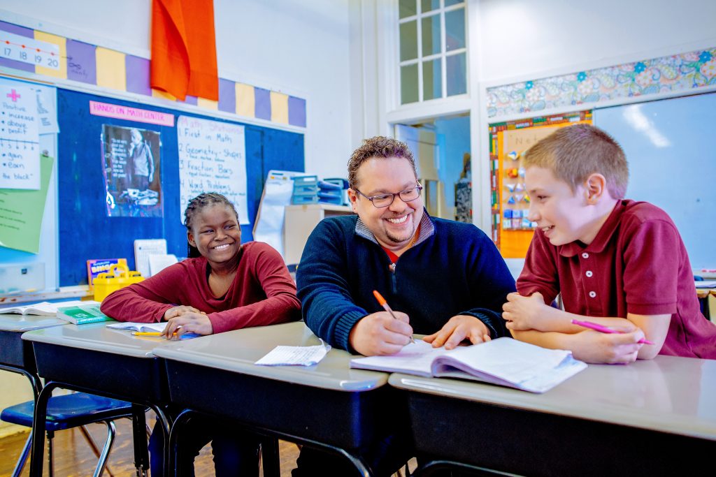 A teacher is sitting at a desk with two students helping them with their