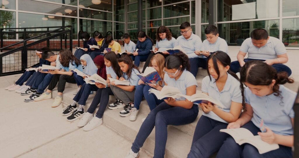 A classroom of students is sitting on the steps outside of school and reading.
