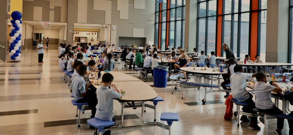 Wide shot of elementary students eating in a cafeteria with windows
