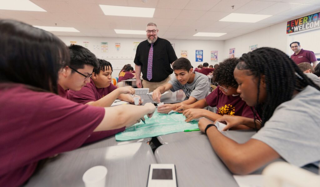 Middle school students around a table working eagerly on a math project, with their teacher encouraging them