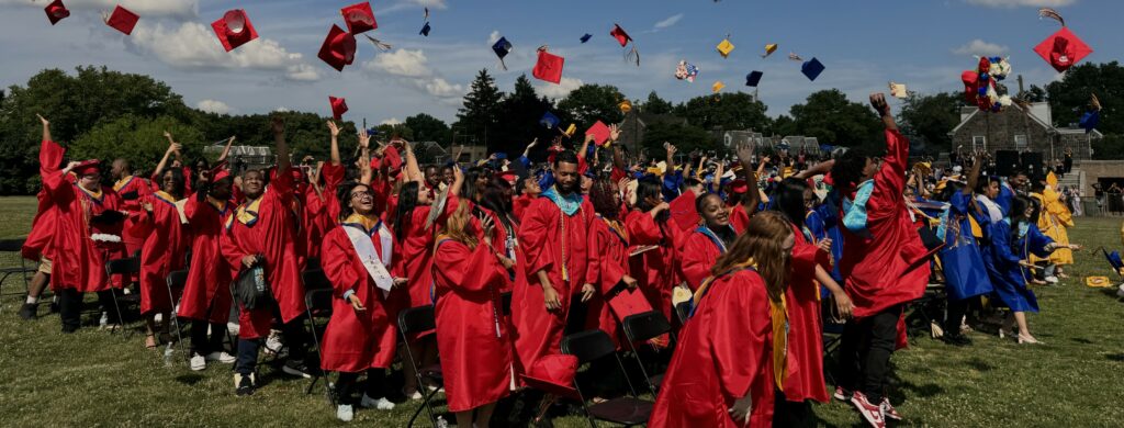 Students at an outdoor high school graduation ceremony, wearing red robes and throwing their caps