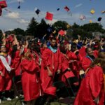 Students at an outdoor high school graduation ceremony, wearing red robes and throwing their caps