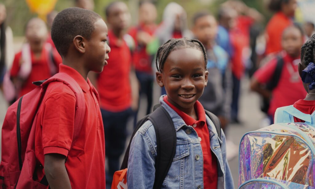 Elementary students in red uniforms standing in line on first day of school, with one student smiling at the camera