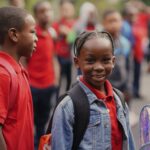 Elementary students in red uniforms standing in line on first day of school, with one student smiling at the camera