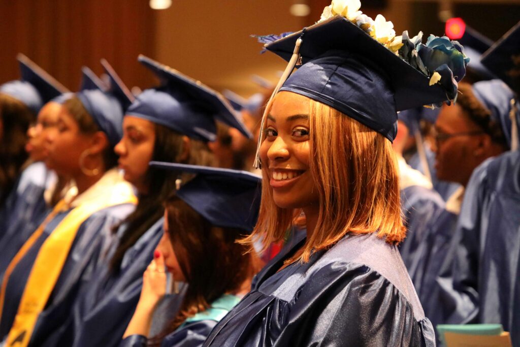 Student in navy cap and gown at graduation, sitting down and looking sideways to smile at camera