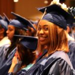 Student in navy cap and gown at graduation, sitting down and looking sideways to smile at camera