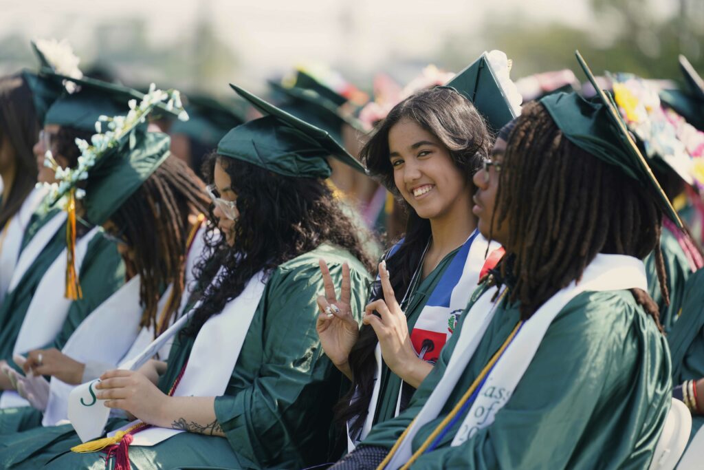 Students sitting in green caps and gowns at high school graduation. One female student is looking at the camera and giving the peace sign.