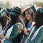 Students sitting in green caps and gowns at high school graduation. One female student is looking at the camera and giving the peace sign.