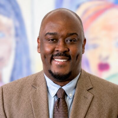 Philadelphia Board of Education member smiling, wearing a brown suit jacket, light blue shirt, and patterned brown tie.
