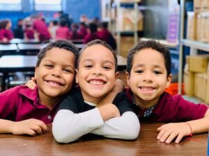 Three children sitting at a table and smiling for the camera while hugging each other.
