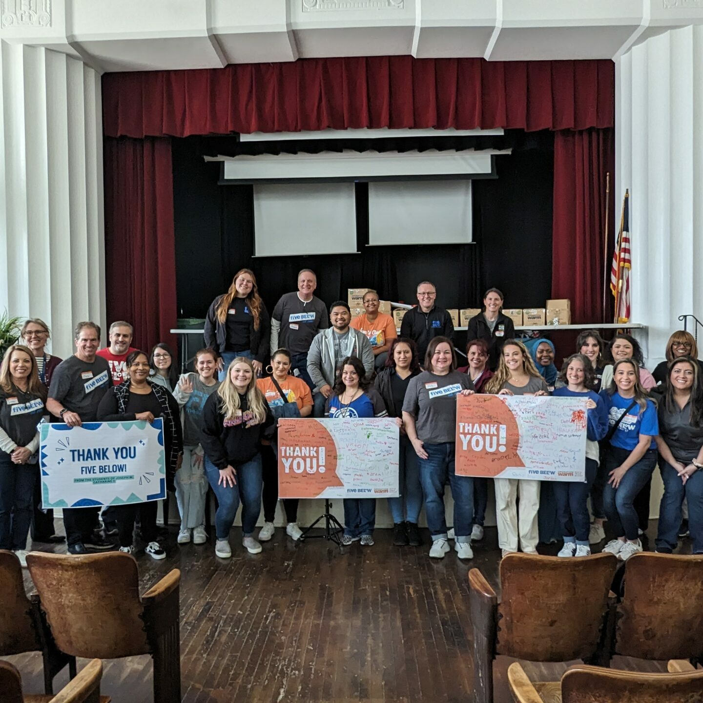 A large group of adults pose on a school auditorium stage holding colorful “Thank You” signs for Five Below, surrounded by flags, flowers, and decorated boxes in the background.