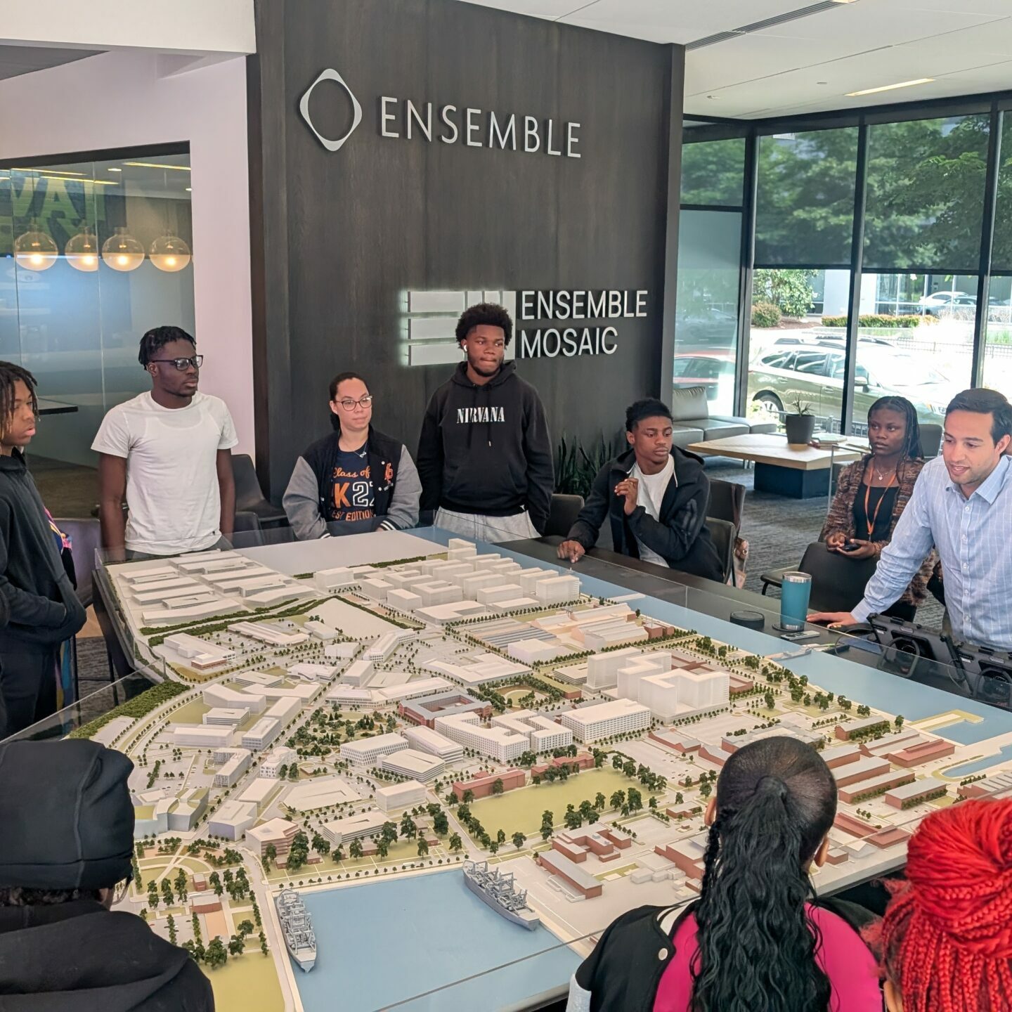 A group of students gather around a large 3D city model at Ensemble Mosaic while listening to a professional explain the development layout in a modern office setting.