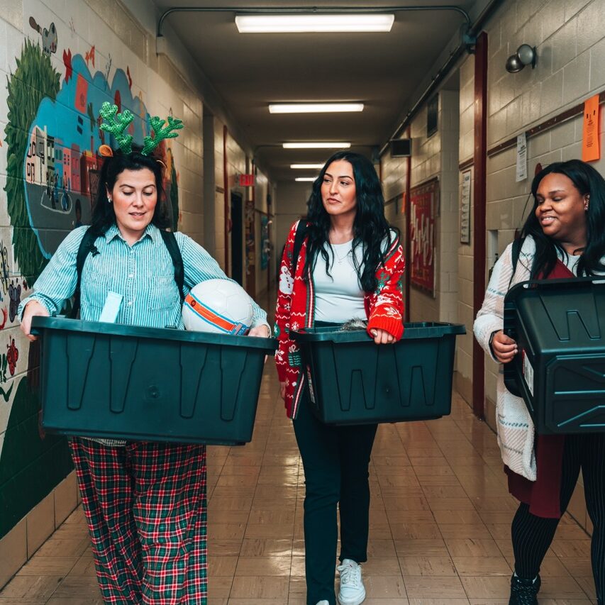 Three women walk down a brightly decorated school hallway carrying large plastic bins. One wears reindeer antlers, another has a red holiday sweater, and they appear to be delivering gifts or supplies.