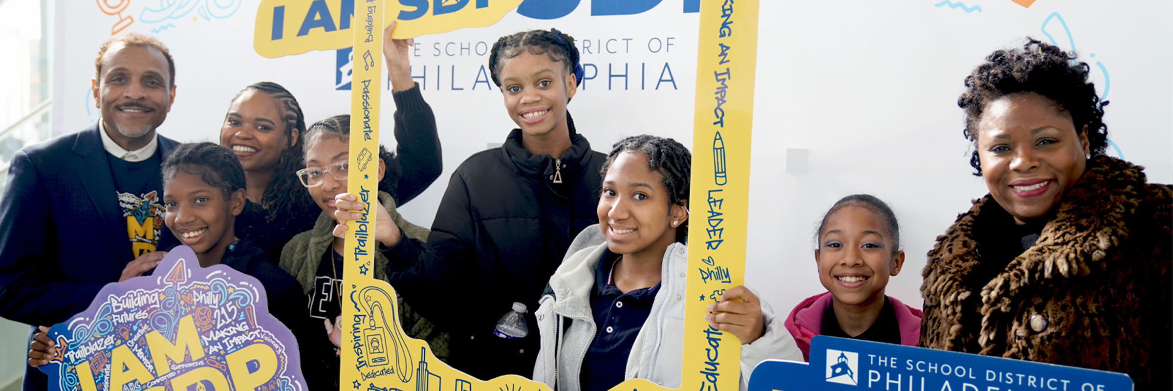 A group of smiling students and School District of Philadelphia staff pose together at an “I AM SDP” photo booth display, holding colorful signs and picture frames that promote pride, leadership, and community.